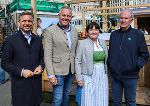 Bildbeschreibung: Eine Frau und drei Männer stehen vor einer Marktkulisse.