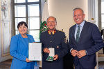 Drei Personen im feierlichen Rahmen. Zwei Personen überreichen eine Urkunde an eine dritte Person.