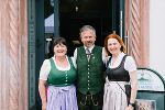 Drei lächelnde Personen in traditioneller Tracht stehen vor dem Eingang eines Gebäudes.