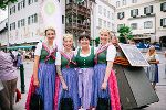 Drei lächelnde Personen in traditioneller Tracht stehen vor dem Eingang eines Gebäudes.